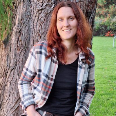 Lektorat J. M. Piel - Bücher, die Vielfalt schaffen. Freies Lektorat für Phantastik. Foto einer weiblich-gelesenen Person, die an einem Baumstamm lehnt und in die Kamera schaut. Sie trägt eine beige Cargohose, ein schwarzes T-Shirt und ein Flanellhemd. Ihre Hände stecken in den Hosentaschen und sie lacht in die Kamera. Ihre roten Haare fallen über ihre Schultern. Im Hintergrund ist eine grüne Wiese. Credit: Wilfried Piel